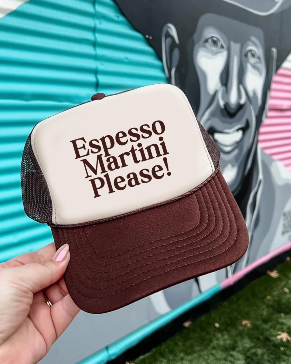 Trucker hat with 'Espresso Martini Please!' text held in front of a colorful mural.
