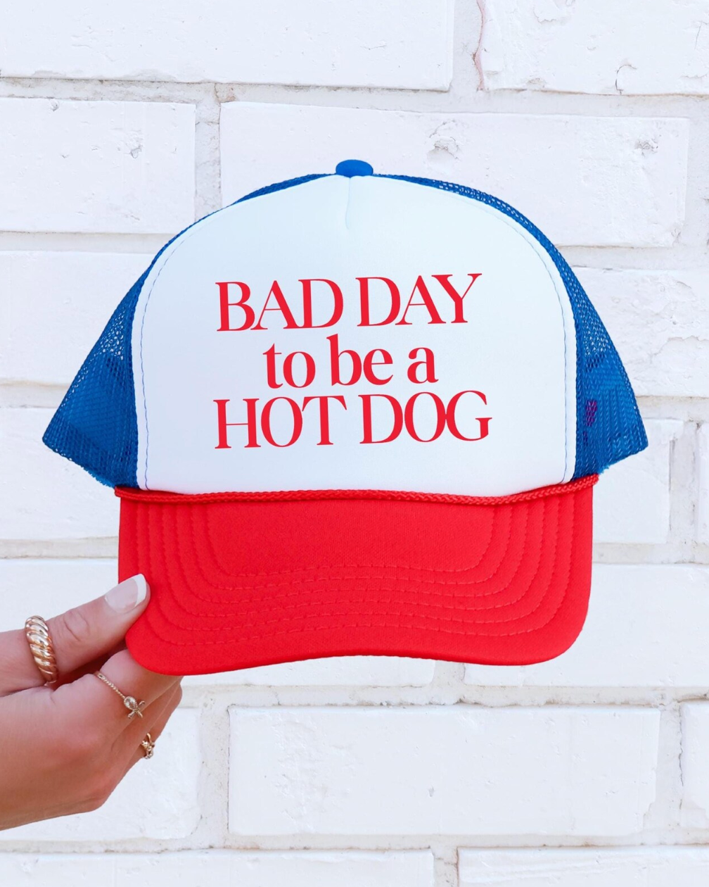 Trucker hat with 'Bad Day to be a Hot Dog' text held against a white brick wall.
