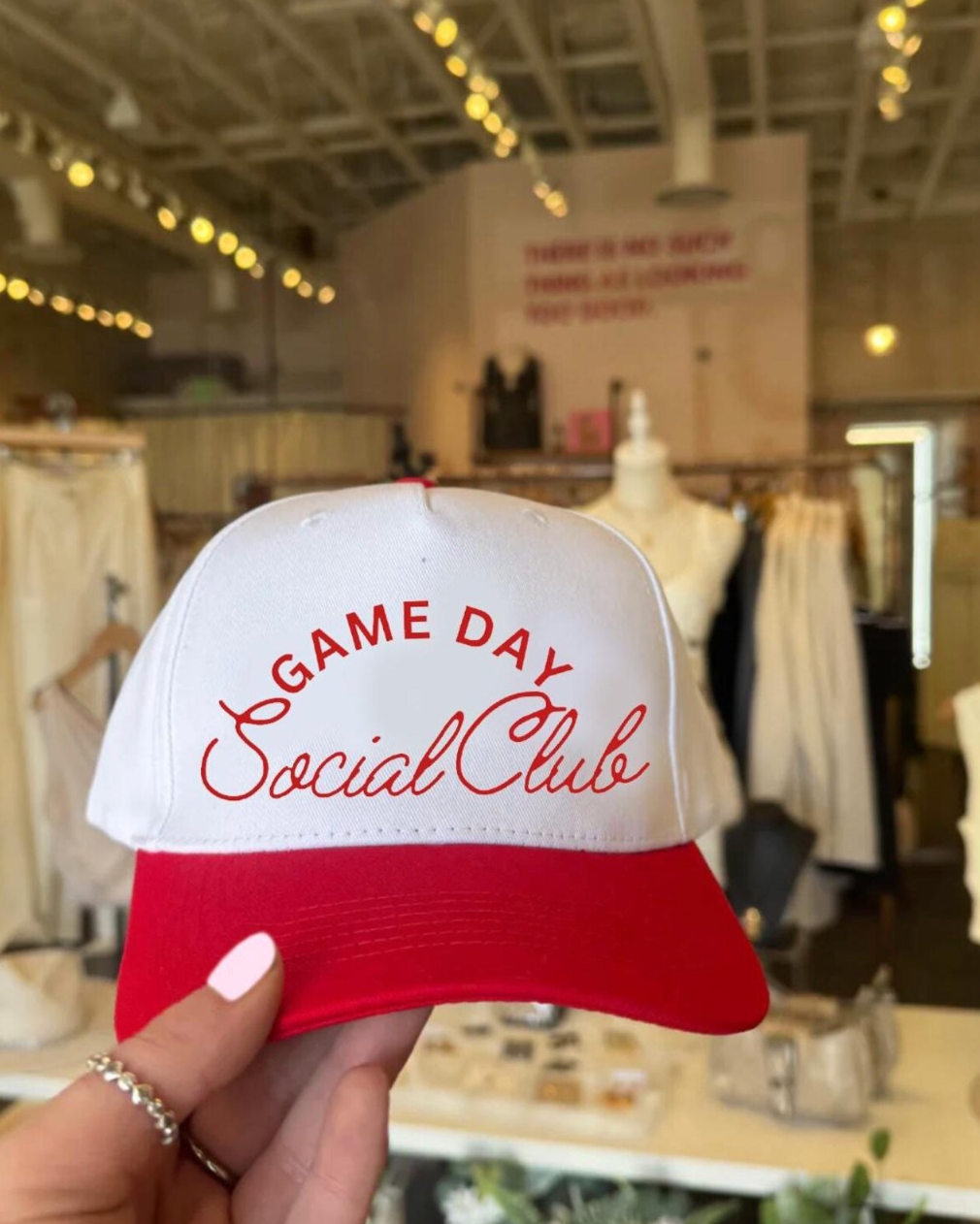 Red and white cap with 'Game Day Social Club' text held in a store setting