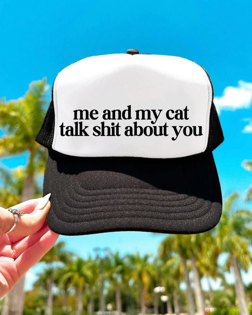 Black and white cap with text held by a hand against a palm tree background