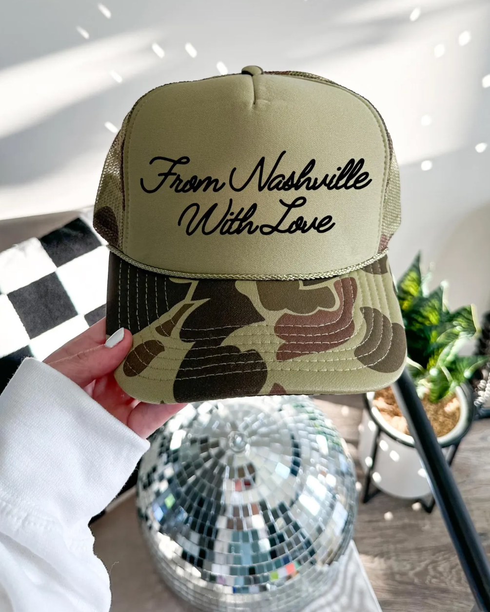 Camouflage cap with 'From Nashville With Love' text held by a person, disco ball in foreground.