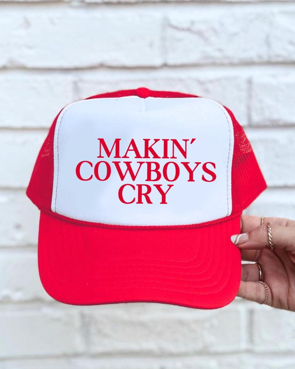 Red and white cap with 'MAKIN' COWBOYS CRY' text held against a white brick wall.