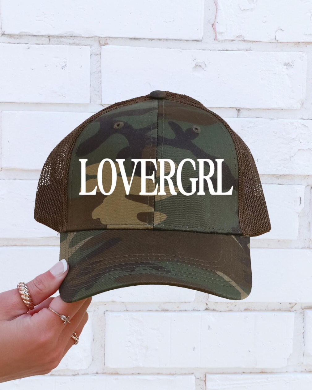Camouflage baseball cap with 'LOVERGRL' text held against a white brick wall.