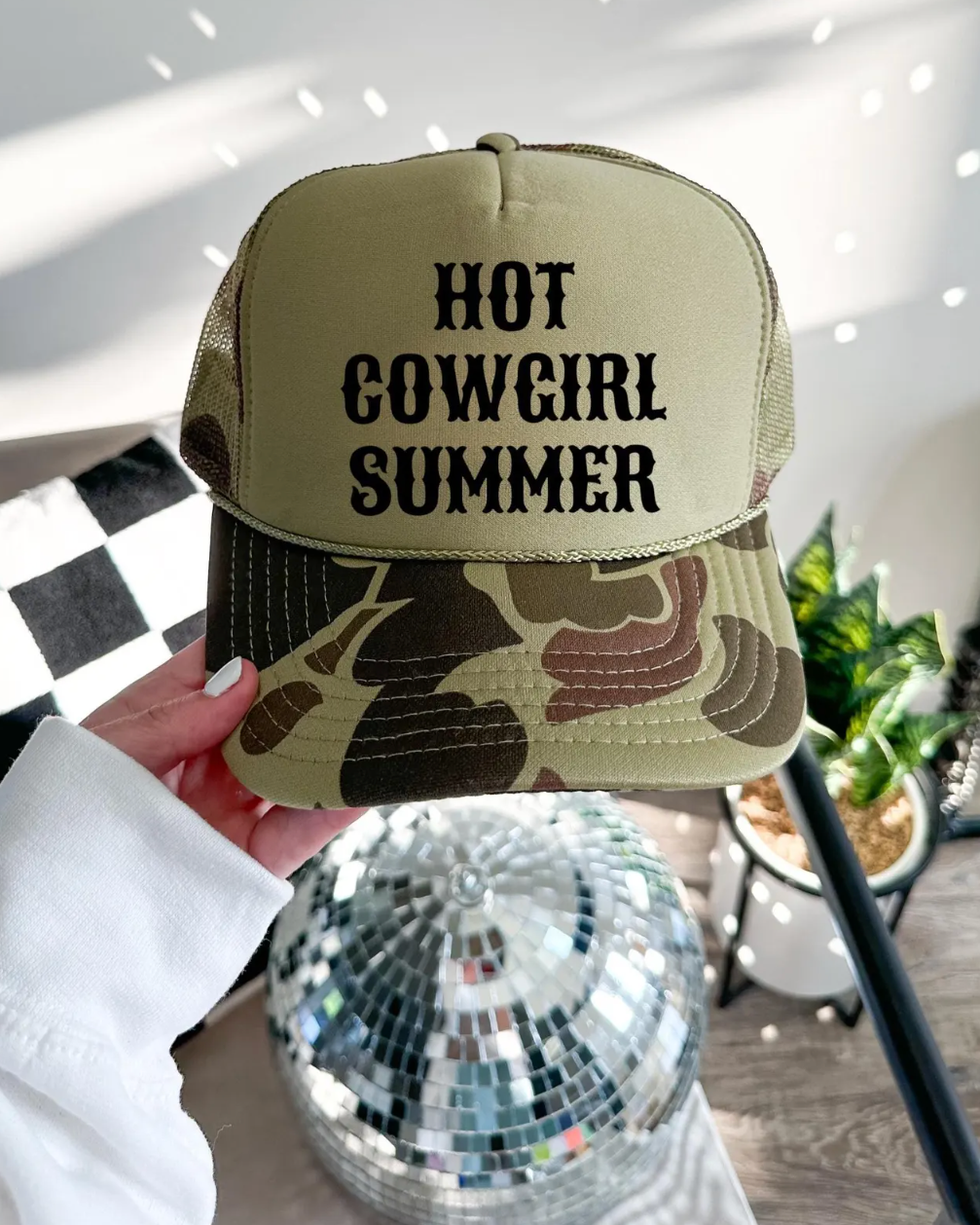 Camouflage cap with 'Hot Cowgirl Summer' text held by a hand in a room with a disco ball and plants.