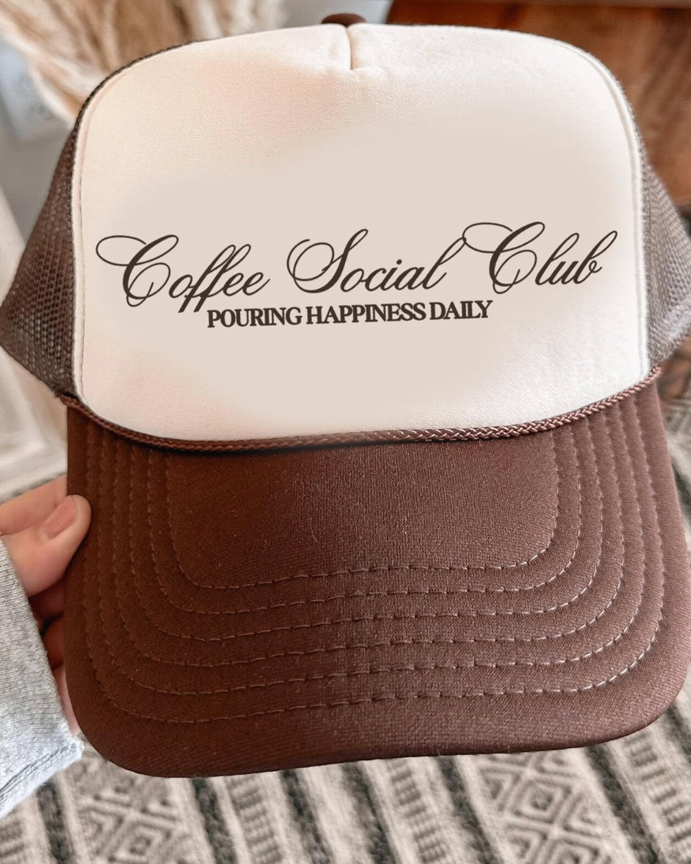 Brown and beige cap with 'Coffee Social Club' text held by a person indoors.