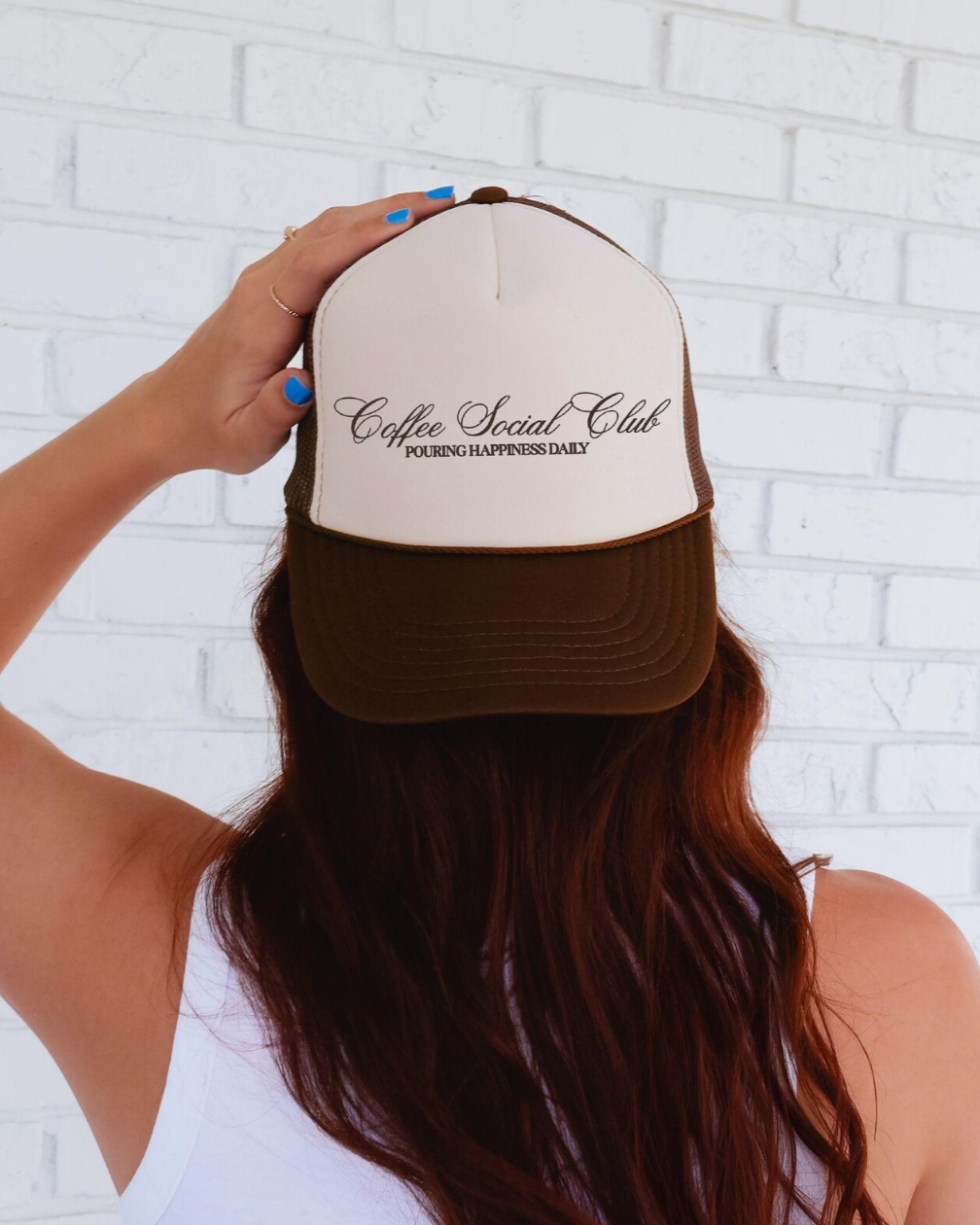Person wearing a brown and white cap with 'Coffee Social Club' text against a white brick wall.