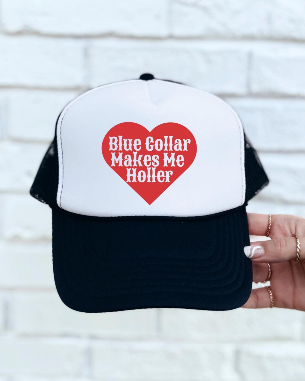 Trucker hat with a red heart and text held against a white brick wall.