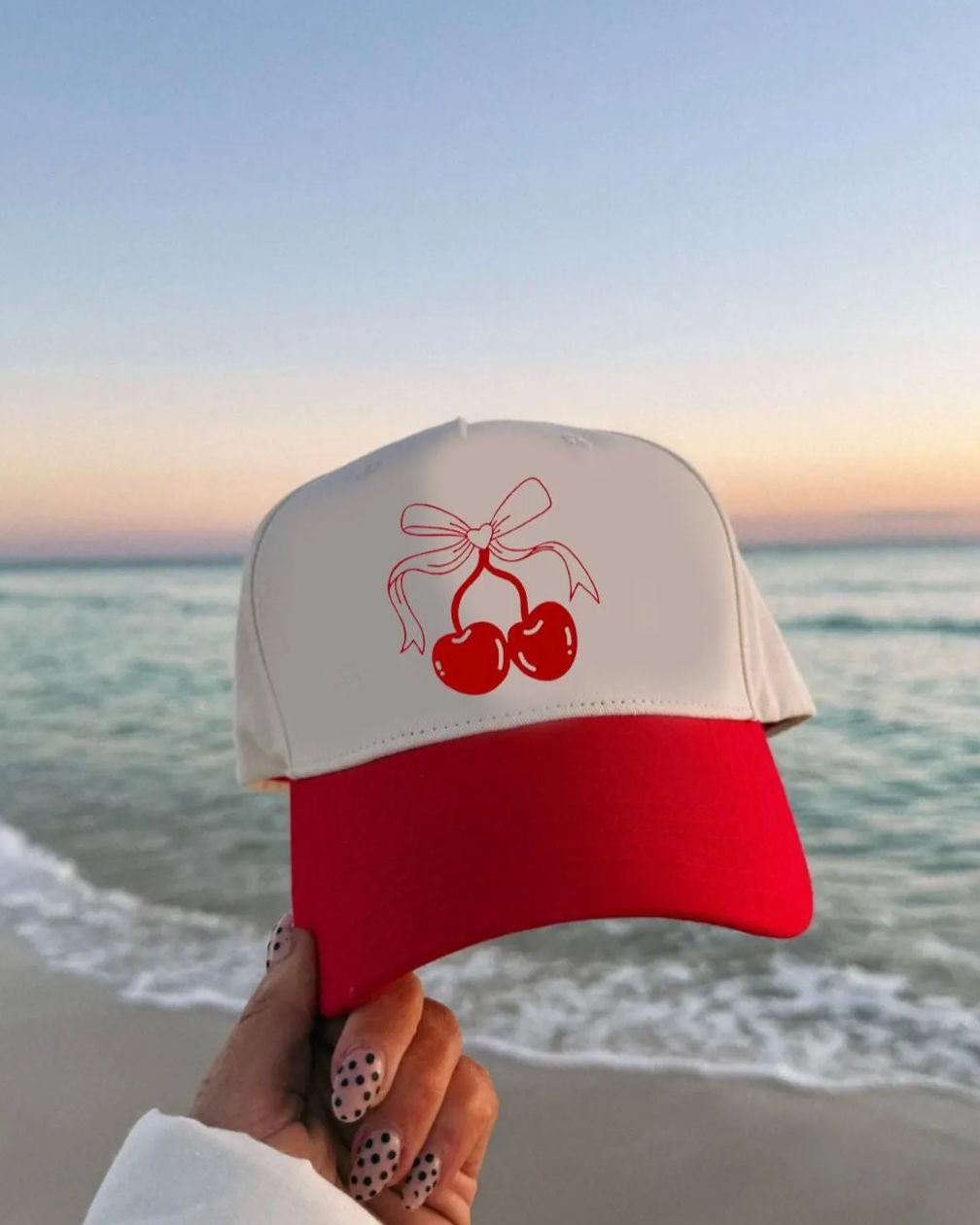 Red and white cap with cherry design held by a hand on a beach