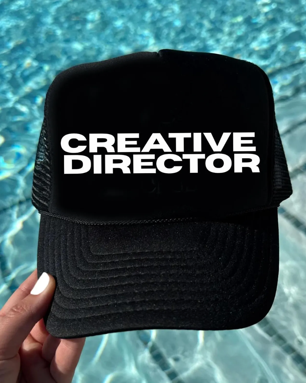 Black cap with 'CREATIVE DIRECTOR' text held in front of a pool.