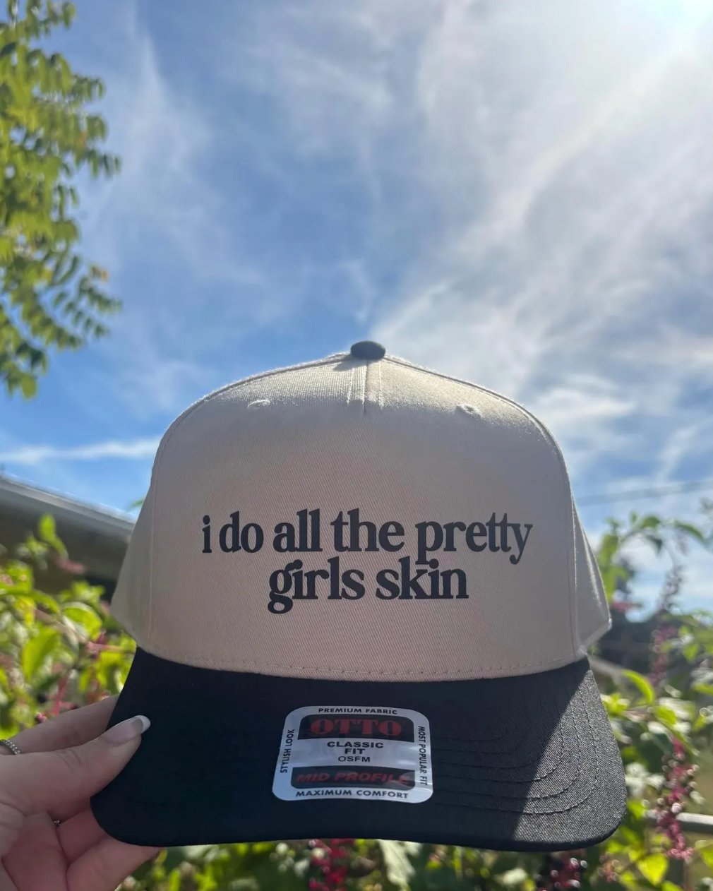 Trucker hat with 'i do all the pretty girls skin' text held against a natural background