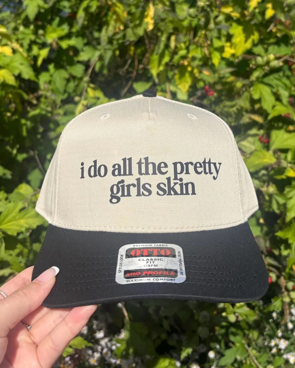 Cap with text 'i do all the pretty girls skin' held in front of green foliage