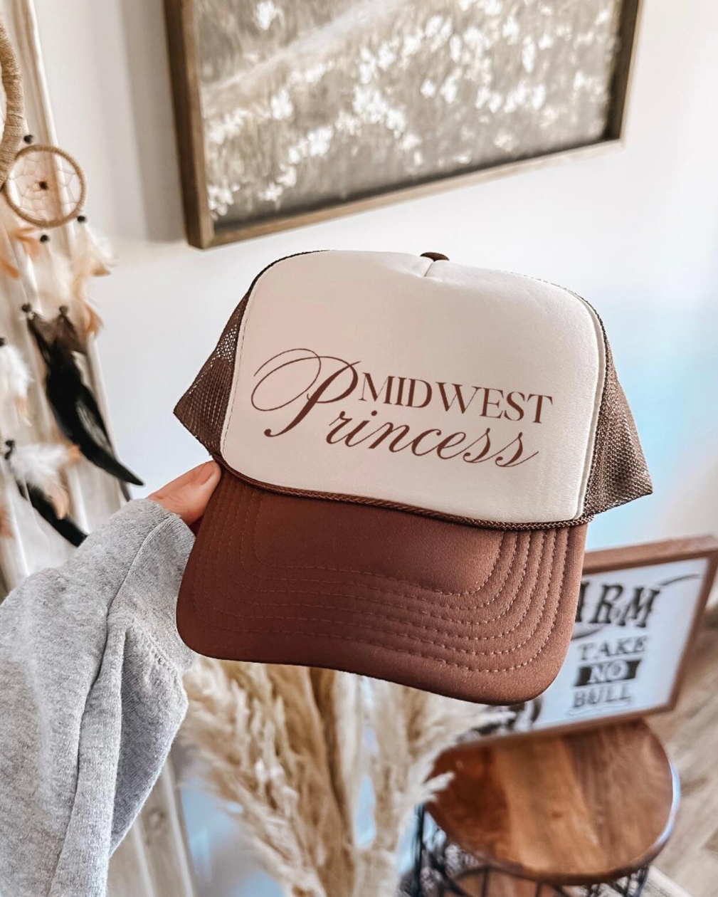 Trucker hat with 'Midwest Princess' text held in a room with decor elements.