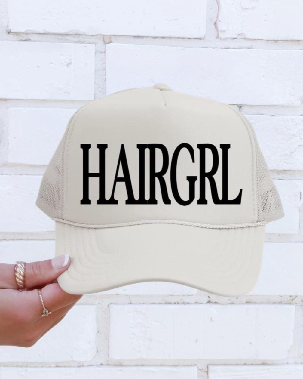 Beige cap with 'HAIRGRL' text held by a hand against a white brick wall.