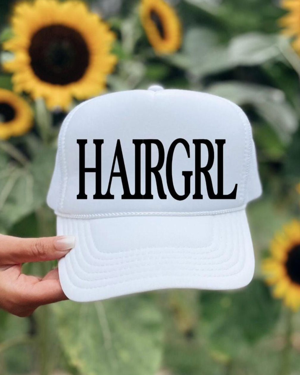 White cap with 'HAIRGRL' text held in front of sunflowers