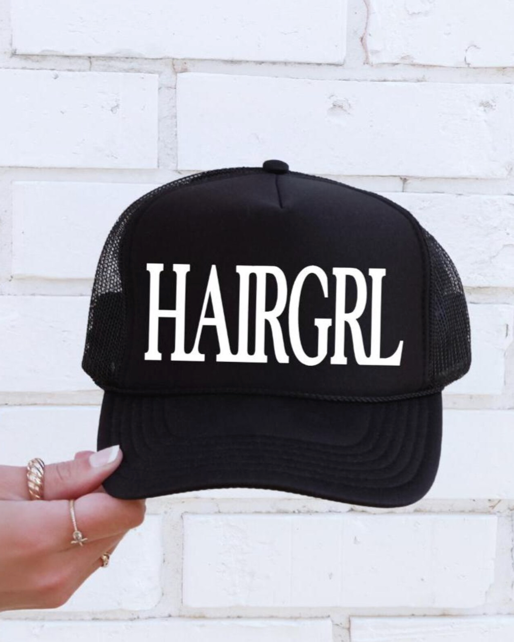 Black cap with 'HAIRGRL' text held against a white brick wall