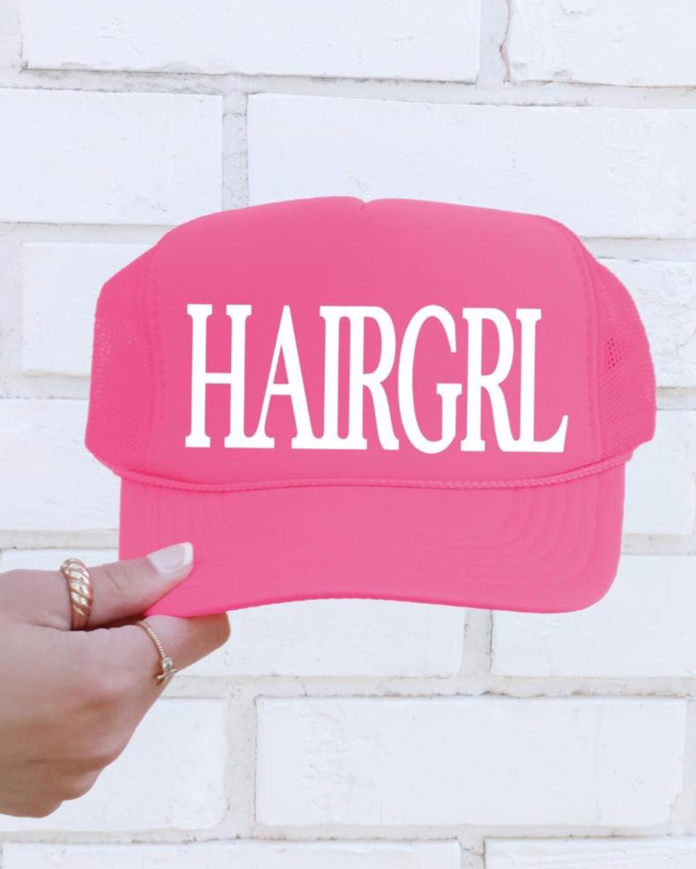 Pink cap with 'HAIRGRL' text held by a hand against a white brick wall.