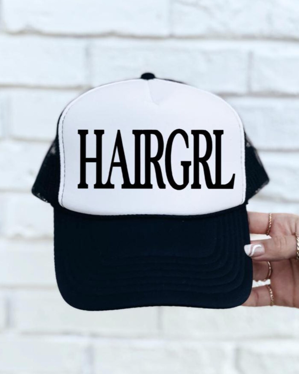 Black and white cap with 'HAIRGRL' text held against a light brick wall.