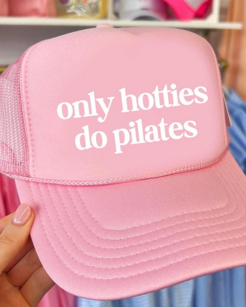 Pink cap with 'only hotties do pilates' text held by a hand in a store setting.