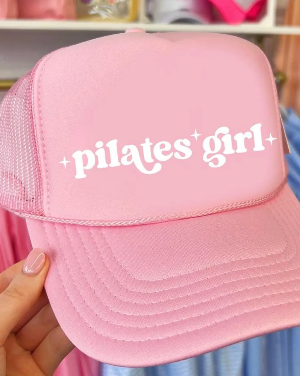 Pink cap with 'pilates girl' text held by a hand in a store setting