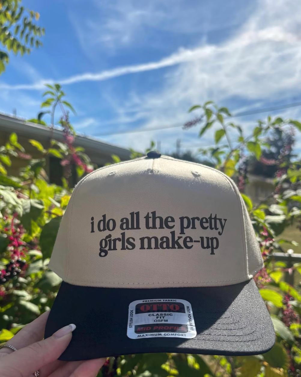 Trucker hat with 'i do all the pretty girls make-up' text held in front of a garden.