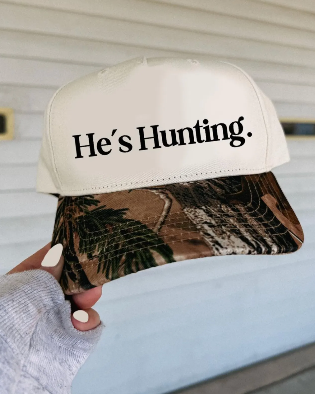 Cap with 'He's Hunting' text on a camouflage bill held by a person.