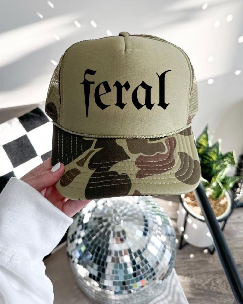 Beige cap with 'feral' text held by a hand, disco ball and plant in the background