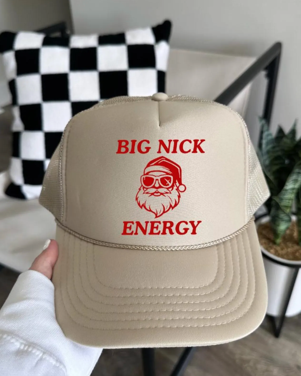 Beige cap with 'BIG NICK ENERGY' text and Santa Claus graphic held by a person indoors.