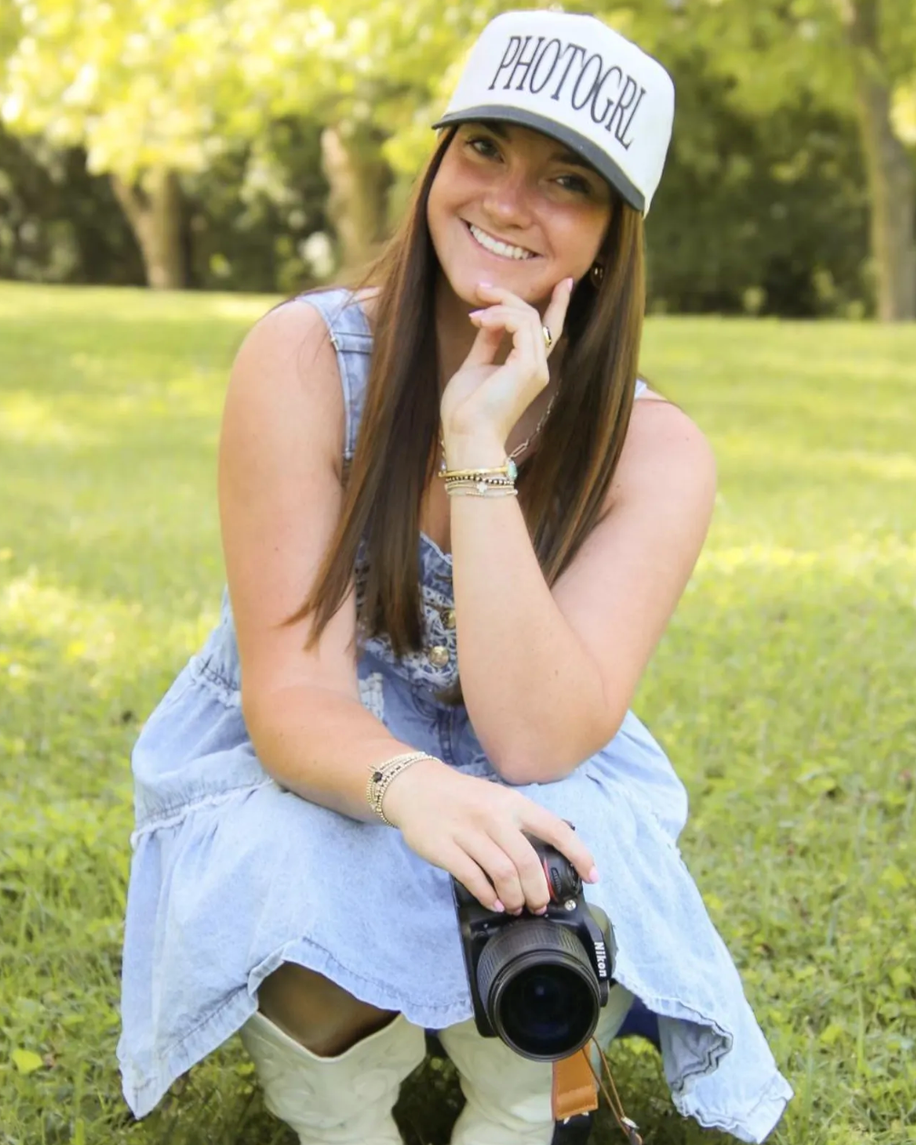 Woman sitting on grass wearing a 'PHOTOGRL' cap and holding a camera.
