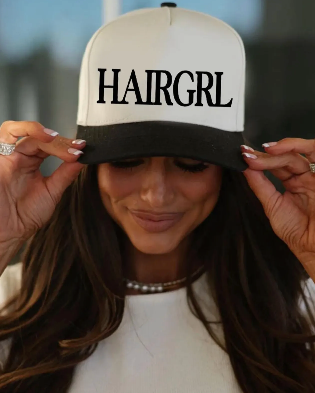 Person wearing a cap with 'HAIRGRL' text, holding it up.