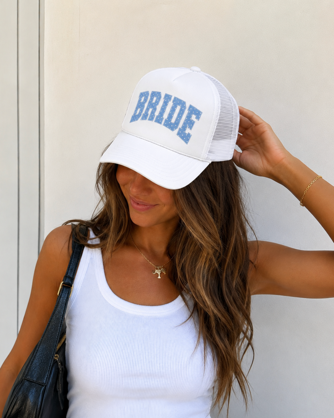 A woman wearing a white trucker-style cap with 'BRIDE' printed in blue block letters on the front, paired with a white ribbed tank top and a black shoulder bag.