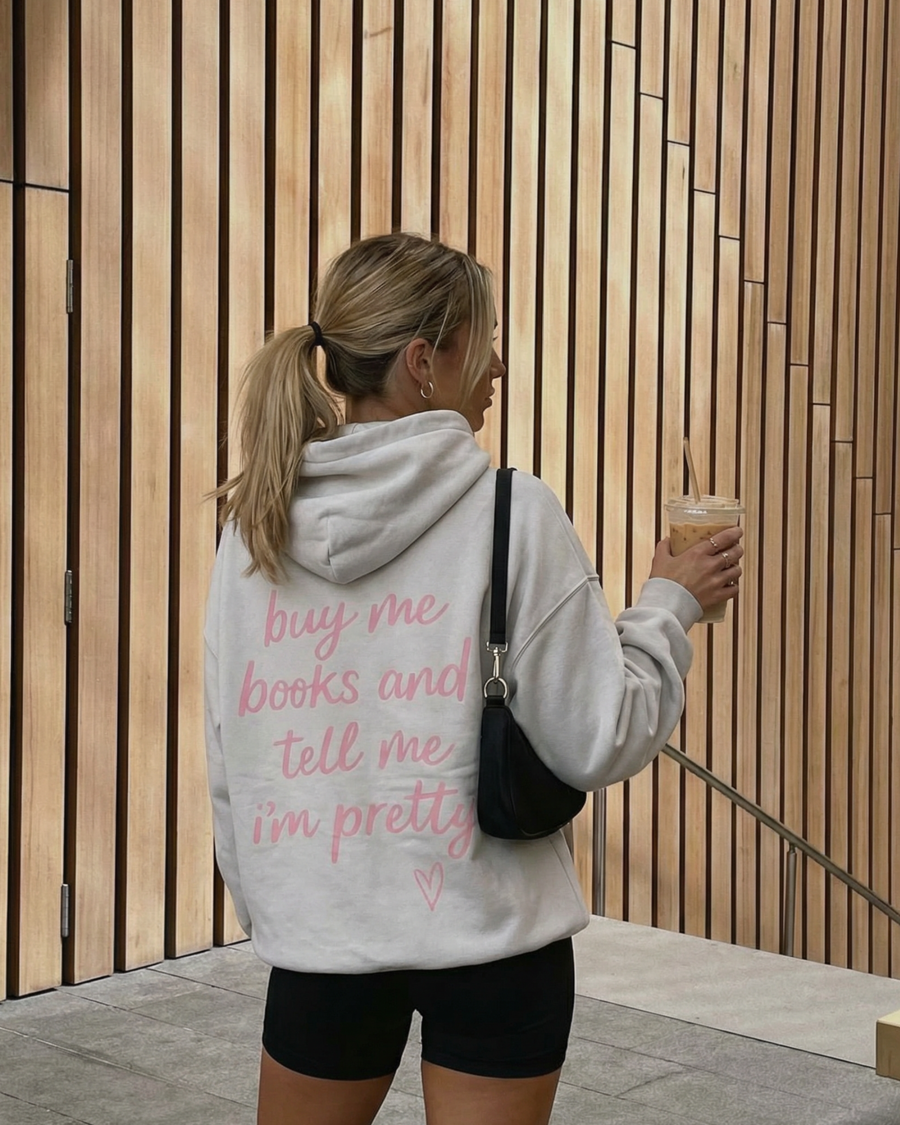 Woman wearing a hoodie with text, holding a drink, standing against a wooden wall.